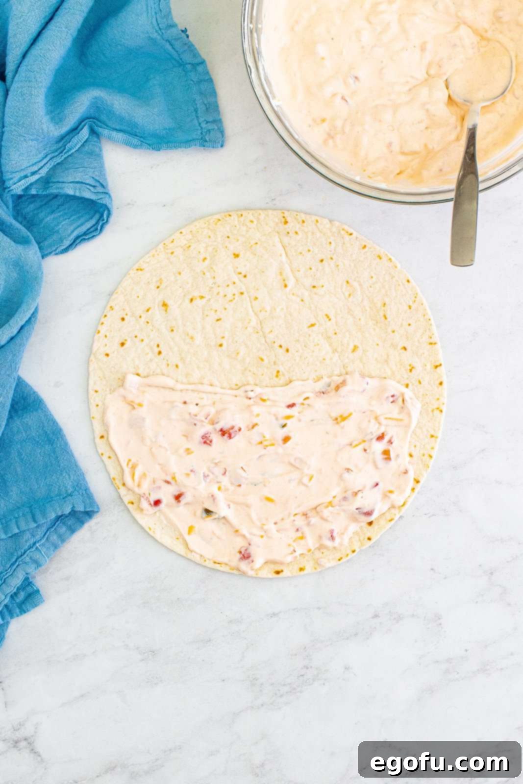A large flour tortilla lies flat on a surface, with a vibrant orange-red salsa cream cheese mixture neatly spread across one half, indicating the next step in preparing a tasty roll-up.
