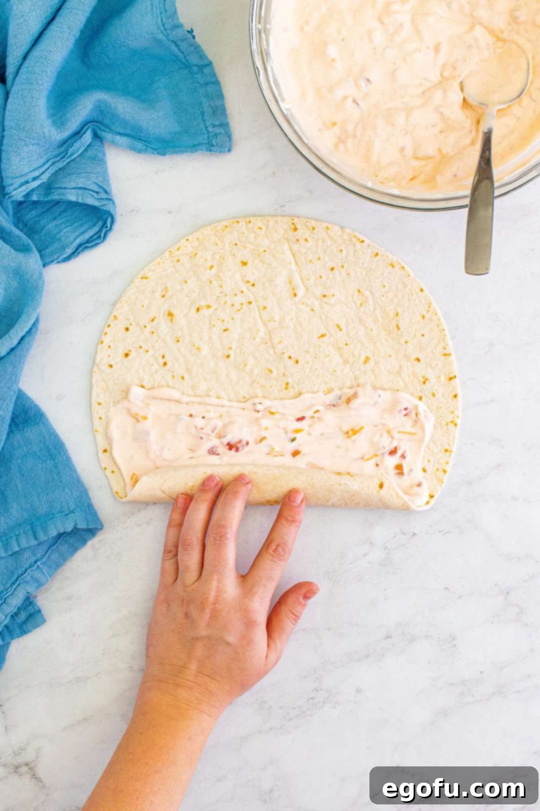 A hand gently but firmly rolls up a flour tortilla filled with a creamy, colorful mixture, demonstrating the technique required to create a tight, uniform cylinder before slicing.