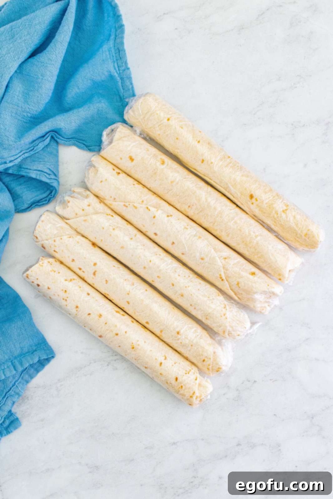 Several rolled flour tortillas, each securely encased in clear plastic wrap, rest side-by-side on a cool, light-colored marble surface, awaiting refrigeration to firm up for slicing.