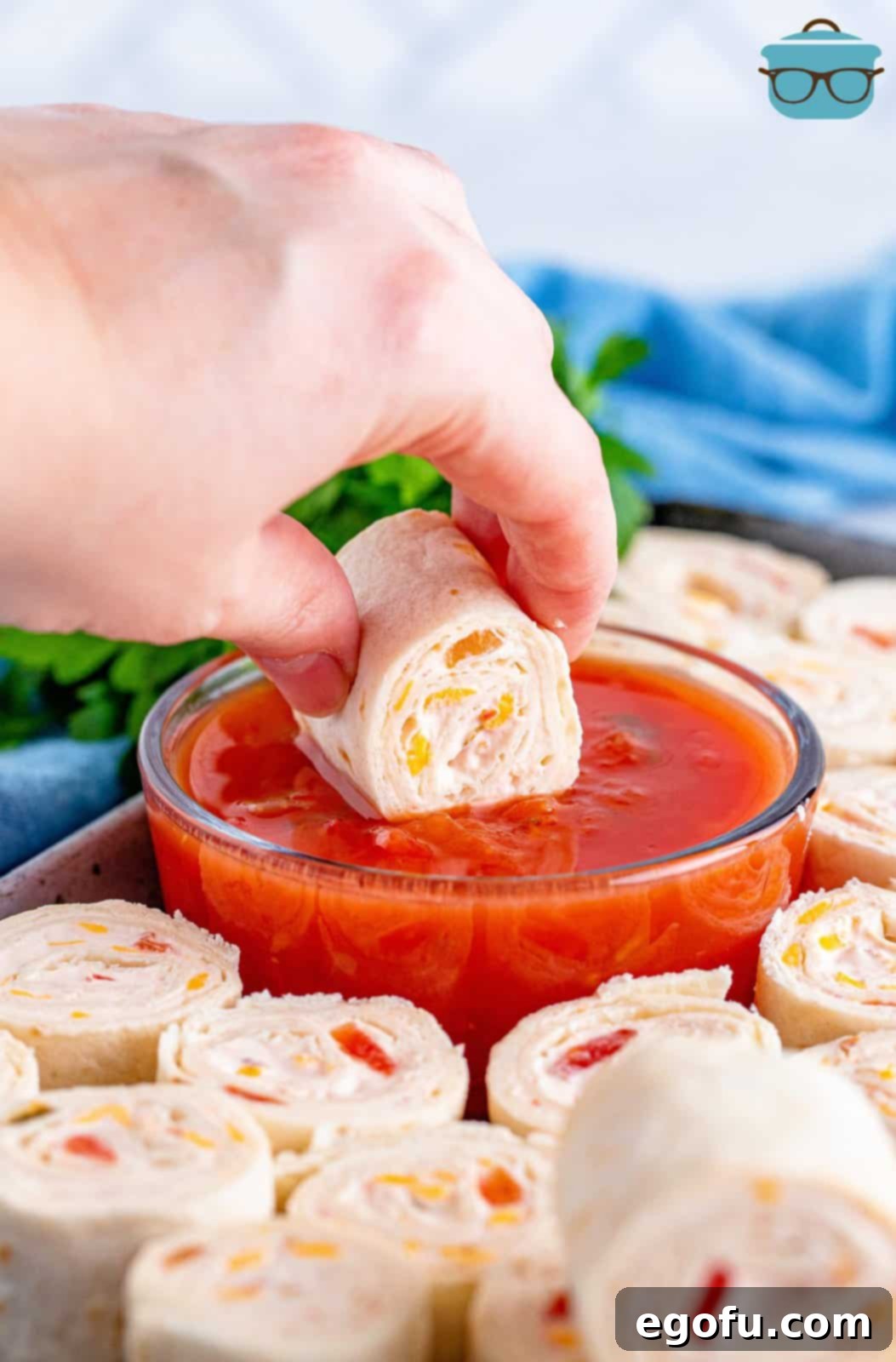 A hand holds a vibrant Fiesta Pinwheel, dipping it into a bowl of bright red salsa, ready to be enjoyed as a refreshing and savory appetizer.