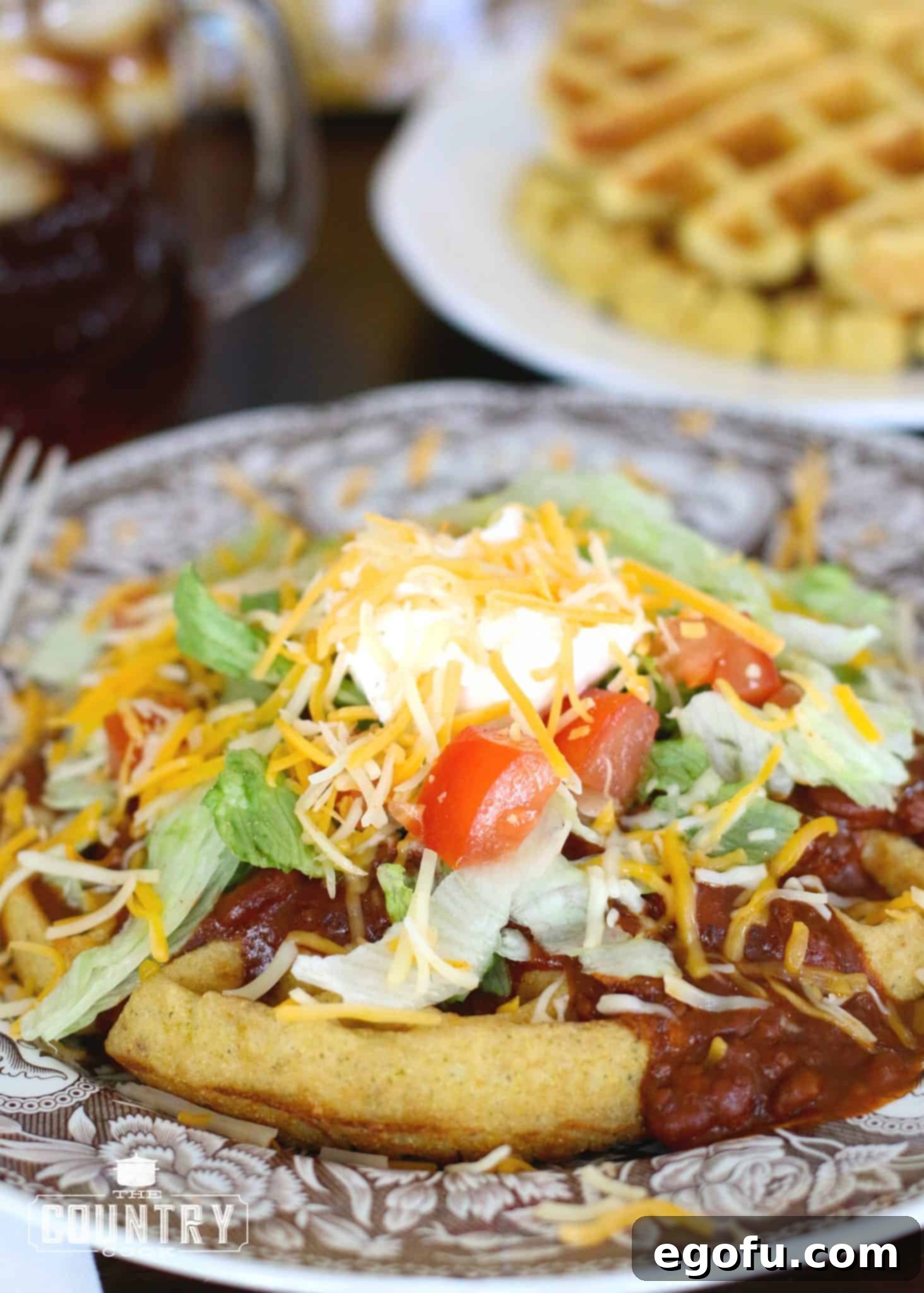 Crispy cornbread waffle generously topped with rich chili, fresh shredded lettuce, melted shredded cheese, a dollop of creamy sour cream, and vibrant diced tomatoes. Served on a rustic brown and white plate.