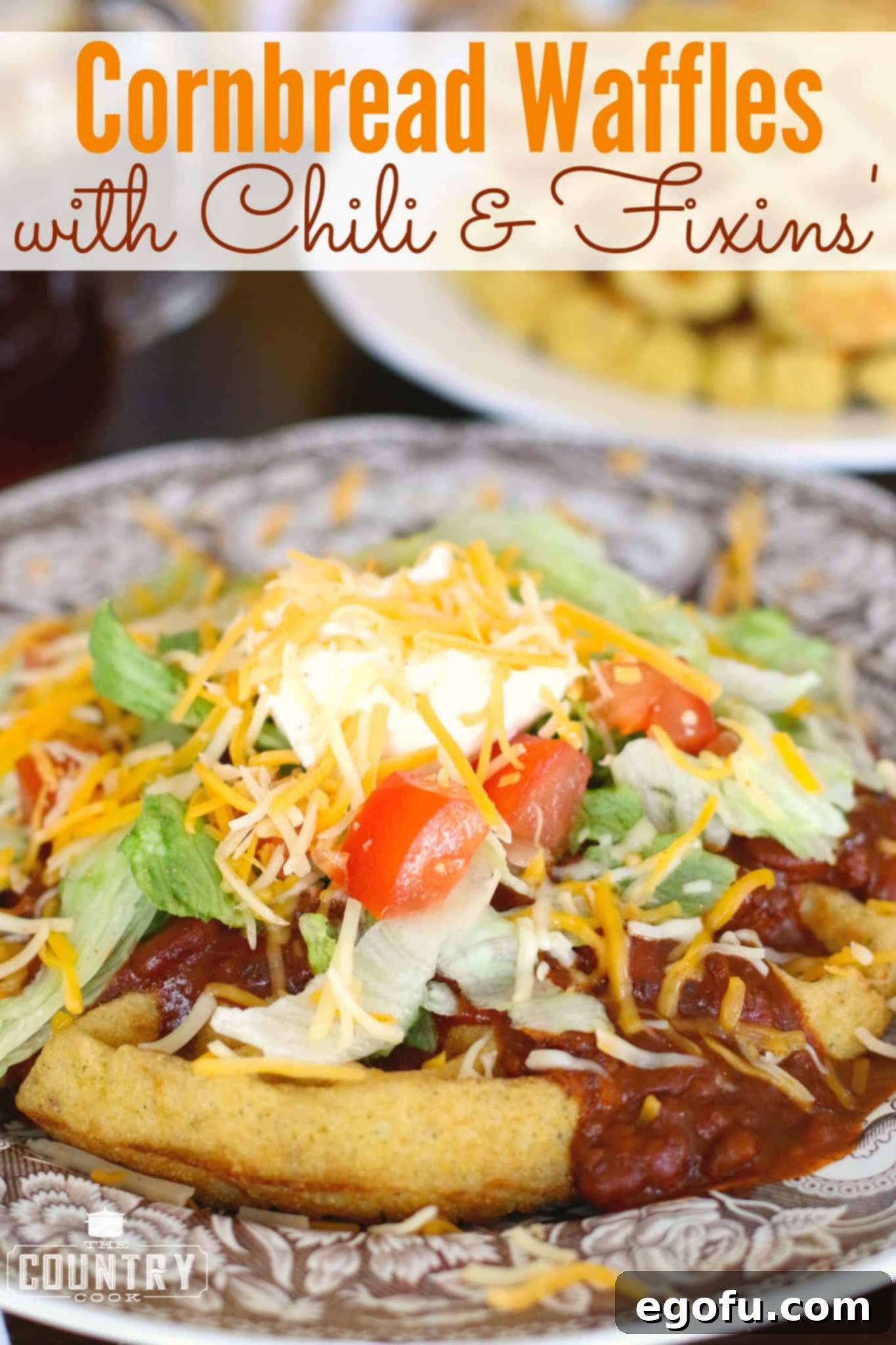 Close-up side view of a delectable cornbread waffle, piled high with savory chili, fresh shredded lettuce, a generous layer of cheese, juicy diced tomatoes, and a dollop of cool sour cream. The vibrant colors and textures are irresistible.