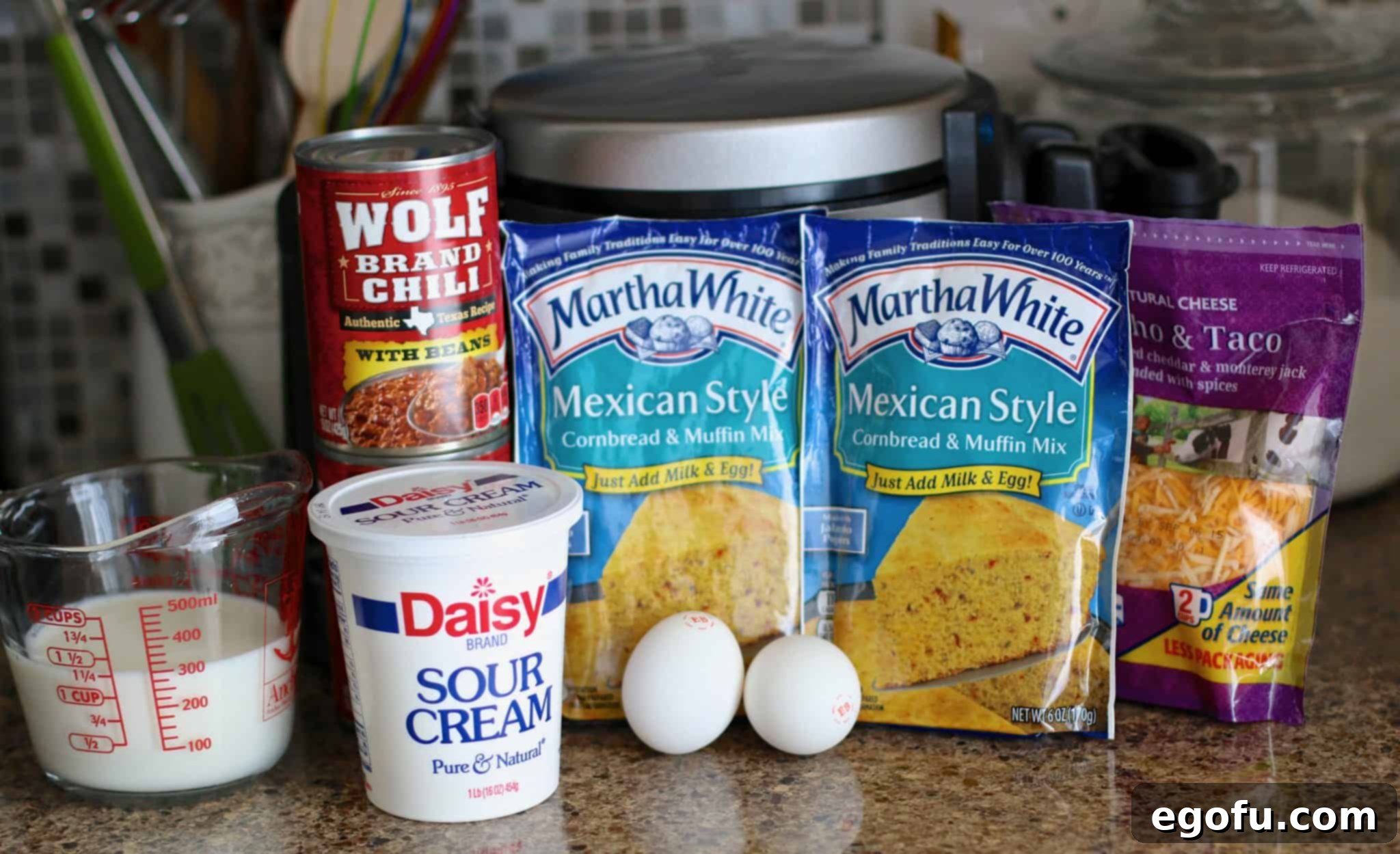 Ingredients laid out for cornbread waffles and chili: two packets of Martha White Mexican Style Cornbread Mix, two cans of Wolf Brand Chili, a bowl of shredded cheddar cheese, two eggs, a tub of sour cream, and milk in a measuring cup.