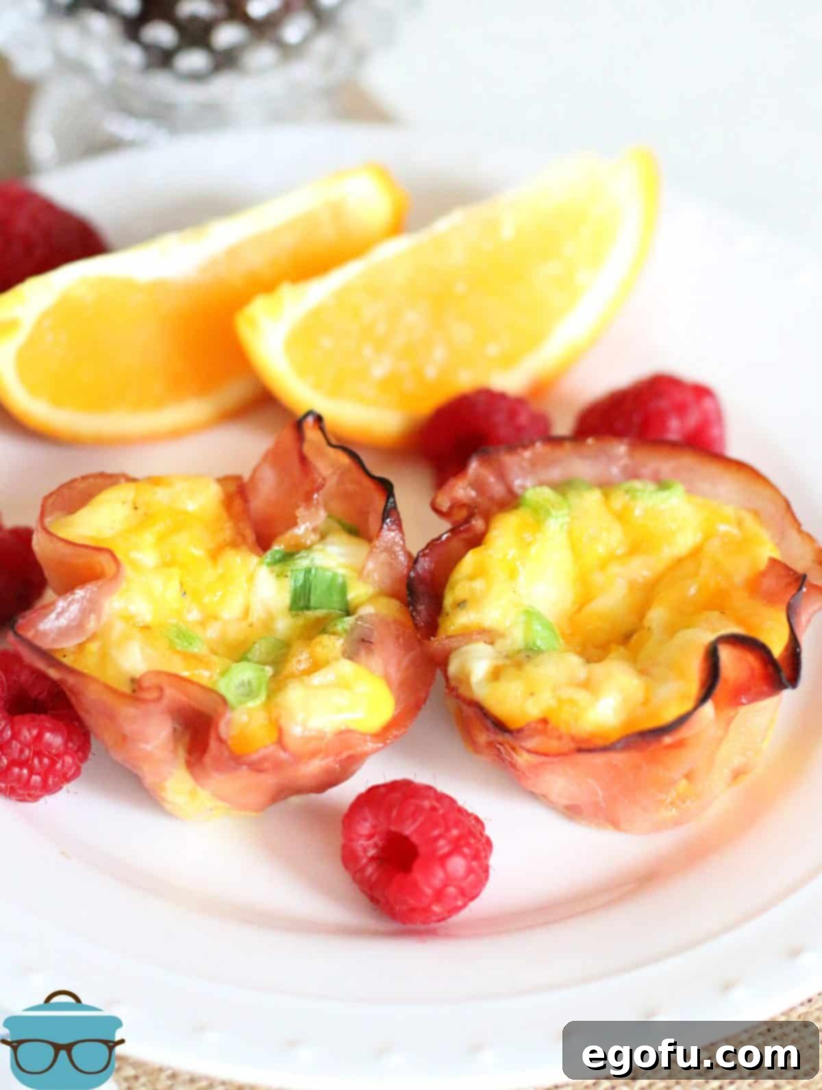Breakfast Ham and Egg Nests 12 two ham and egg cups shown on a white plate with fresh raspberries and sliced oranges.