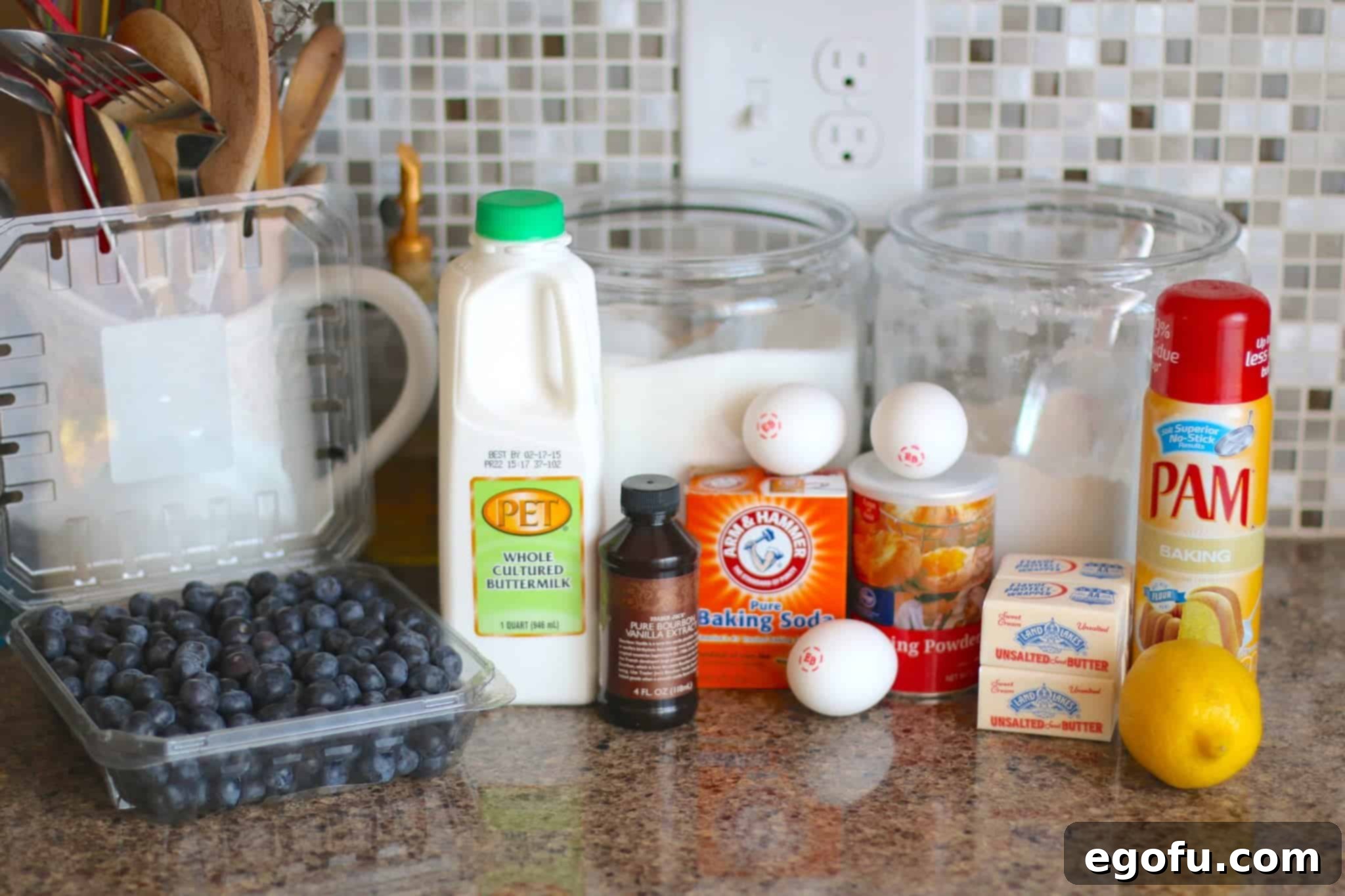 A flat lay image displaying the fresh and high-quality ingredients for the blueberry muffin cake, including vibrant blueberries, creamy buttermilk, butter, vanilla extract, lemon extract, baking soda, baking powder, and fresh eggs.