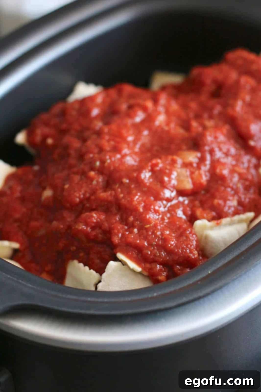 Frozen ravioli in an oval slow cooker topped with a vibrant red sauce, before stirring.