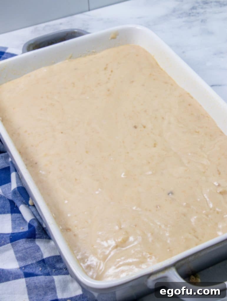 The second half of the banana cake batter being poured and spread gently over the first layer of crumb mixture in the baking dish.