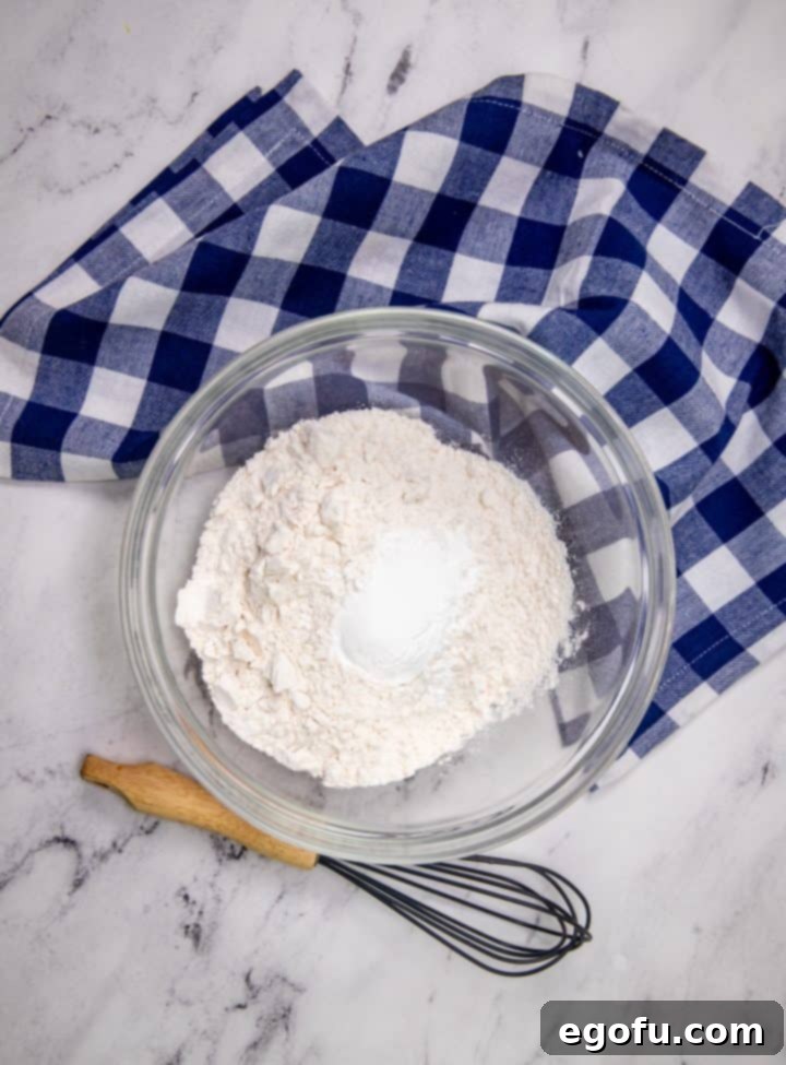 Close-up of a clear glass bowl containing all-purpose flour, baking powder, and salt, with a metal whisk resting beside it, ready for mixing dry ingredients.