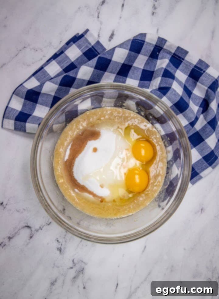 Mashed ripe bananas mixed with melted butter, granulated sugar, and eggs in a clear glass mixing bowl, accompanied by a blue and white checkered tea towel.