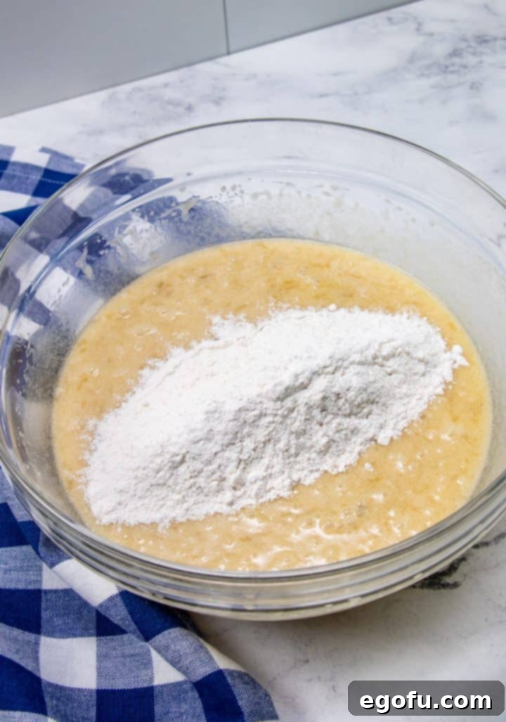 The flour mixture being slowly stirred into the mashed banana and wet ingredients in a clear mixing bowl, creating a smooth cake batter.