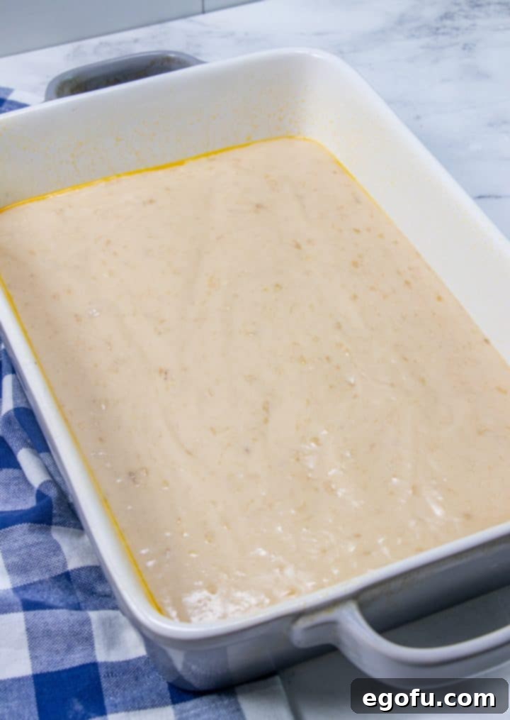 Half of the banana cake batter smoothly spread across the bottom of a prepared 9x13 inch baking dish, forming the first layer.