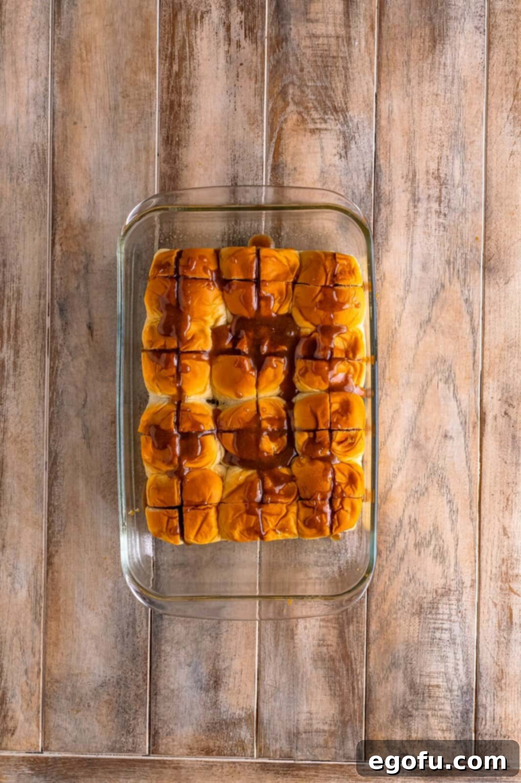 Warm cinnamon glaze being spooned into the opened slices of the Hawaiian sweet rolls in the baking dish, ensuring even distribution.