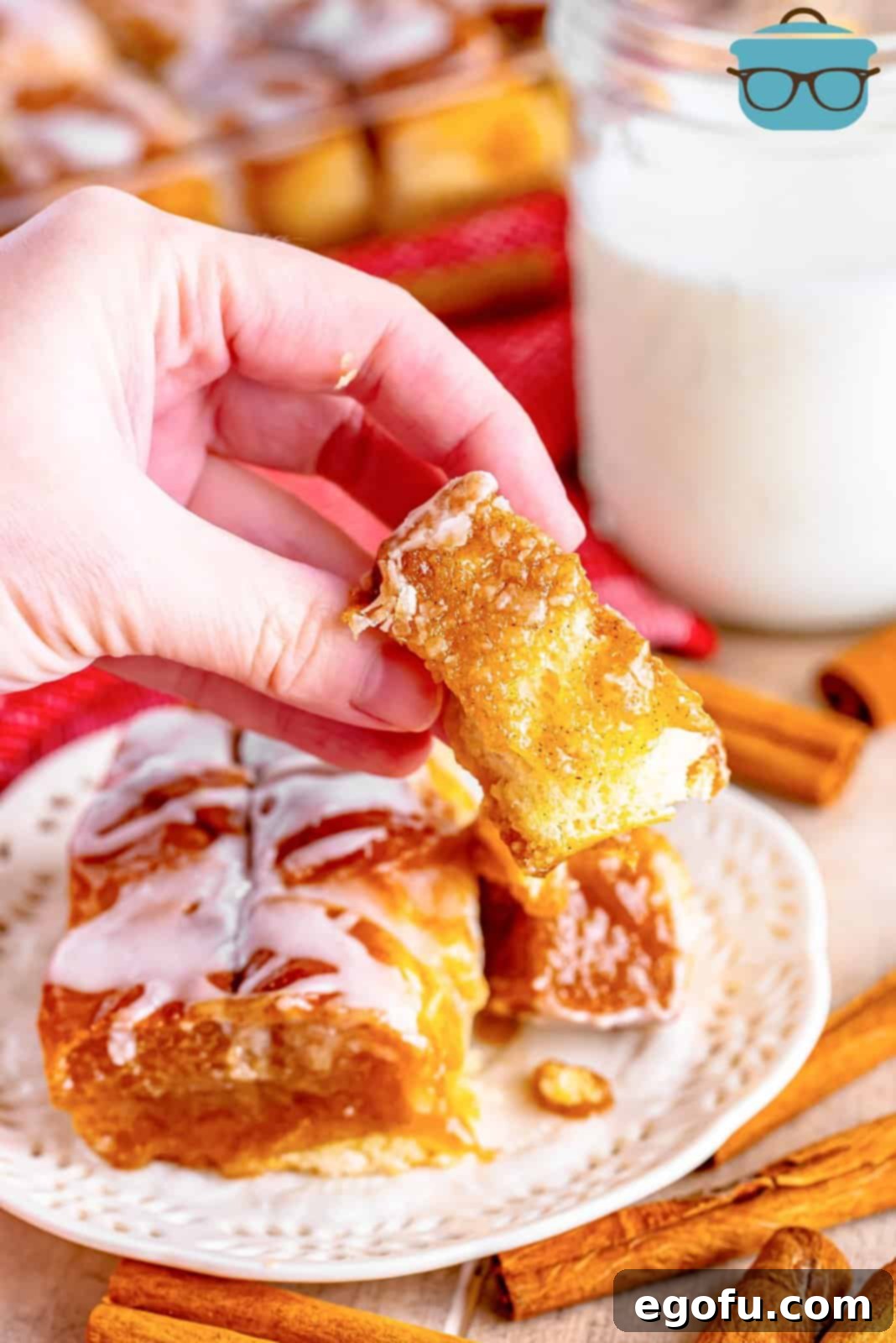 A hand presenting a perfectly pulled-apart, glazed piece of cinnamon bread in front of a blurred background of a plate and a glass of milk, inviting a taste.