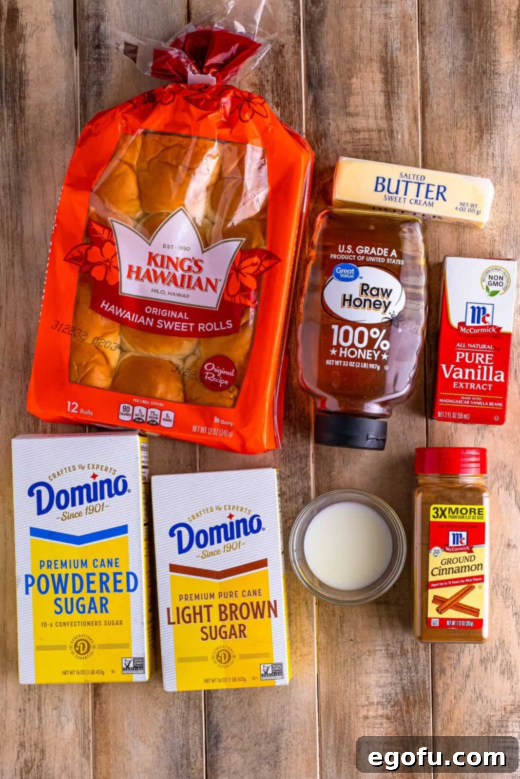 A beautifully arranged flat lay of all the essential ingredients for making Hawaiian sweet rolls, including a package of sweet rolls, light brown sugar, powdered sugar, ground cinnamon, sticks of butter, vanilla extract, and a small jug of milk, ready for baking.