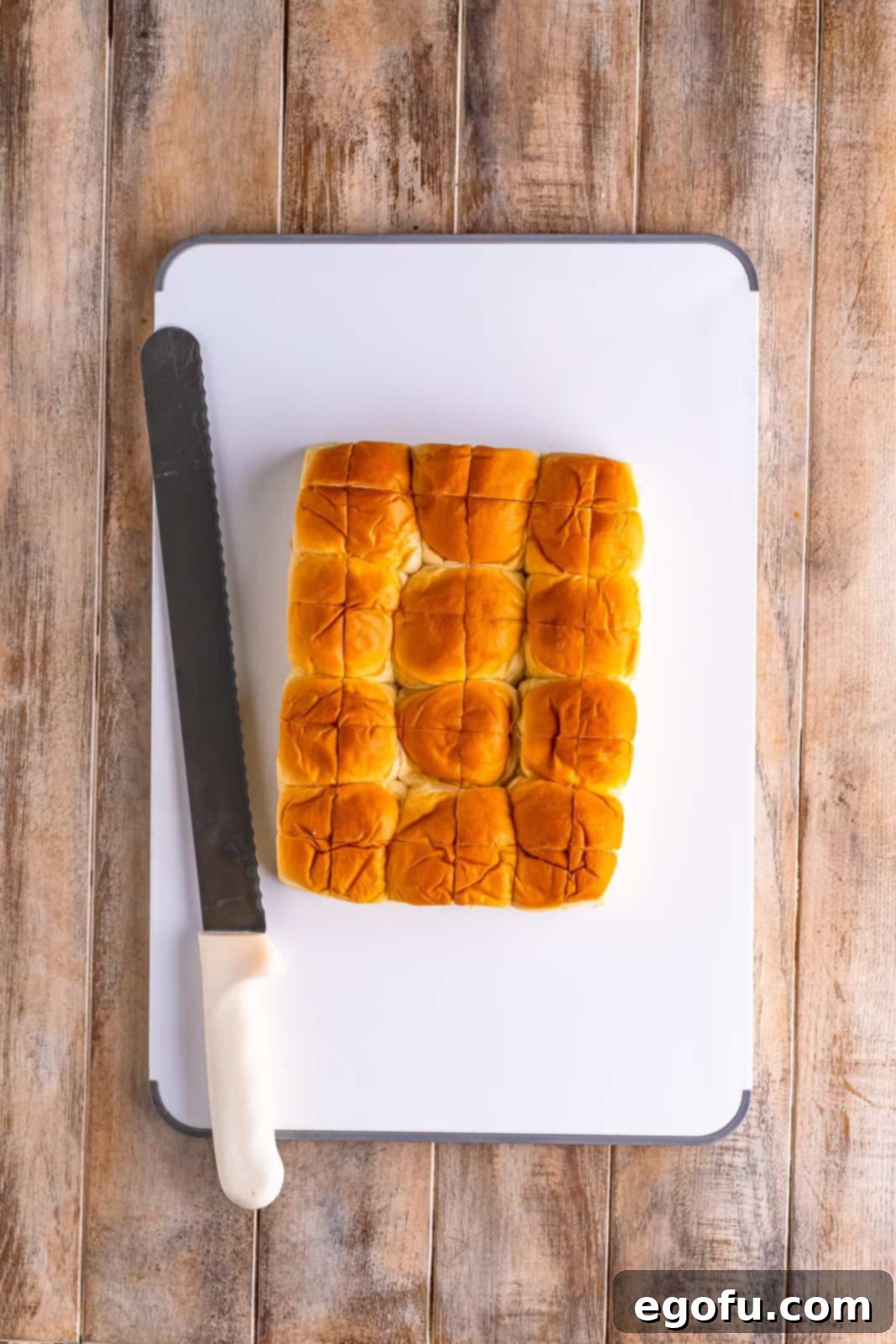 A serrated knife completing the cross-hatch cuts on the King's Hawaiian sweet rolls, showing the preparation for the pull-apart bread.
