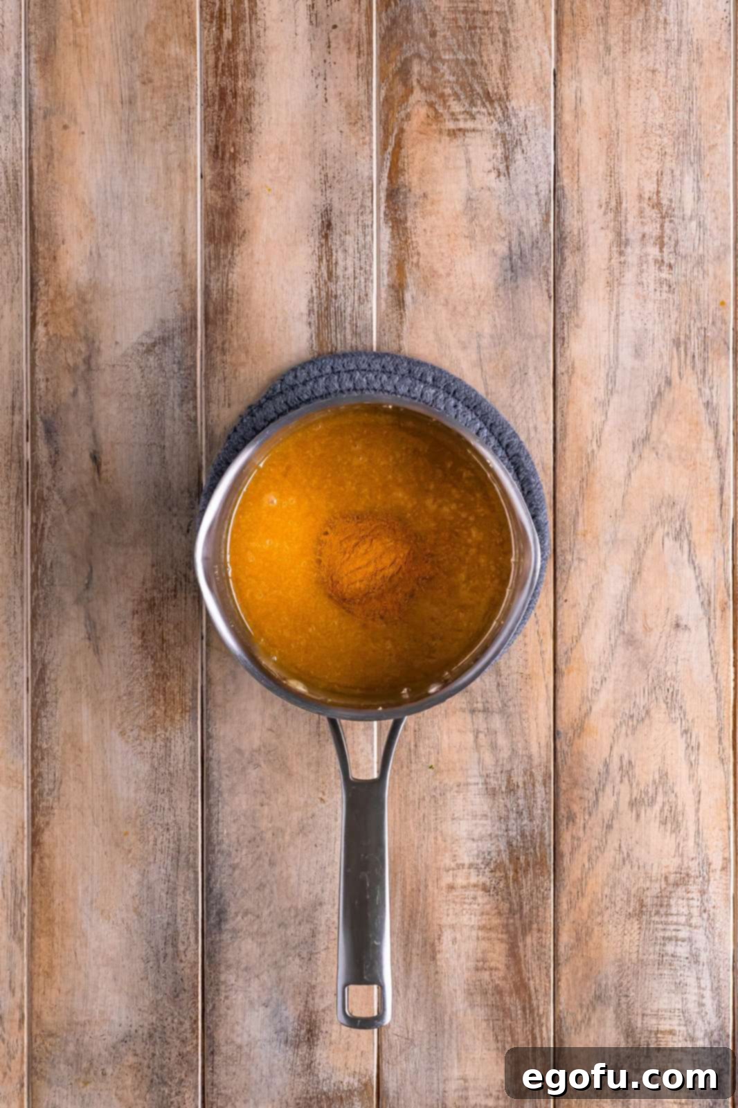 Ground cinnamon being stirred into the warm, melted glaze mixture in a saucepan, blending into a fragrant cinnamon-sugar syrup.