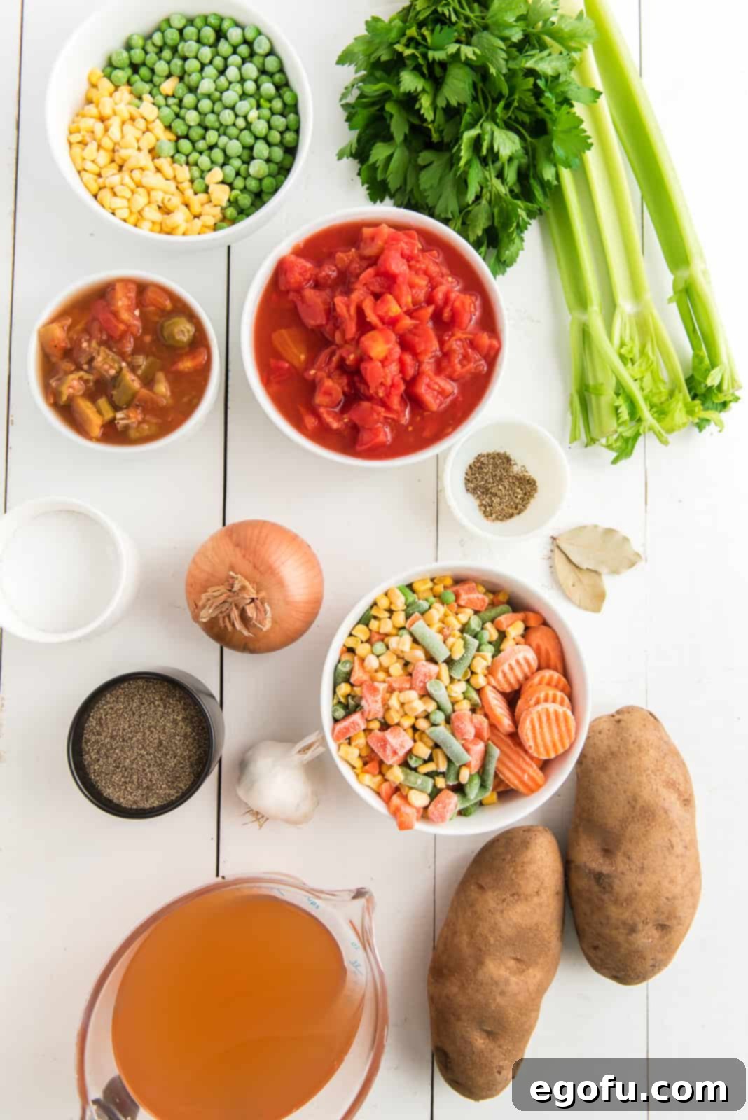 One Pot Comfort Vegetable Soup 4 A flat lay photograph displaying all the fresh and frozen ingredients needed for Crock Pot Vegetable Soup: onion, celery, olive oil, garlic, frozen mixed vegetables, frozen sliced carrots, frozen peas, frozen corn, diced tomatoes, tomatoes and okra, vegetable broth, potato, bay leaves, salt, pepper, dried basil, dried oregano and a sprig of parsley.