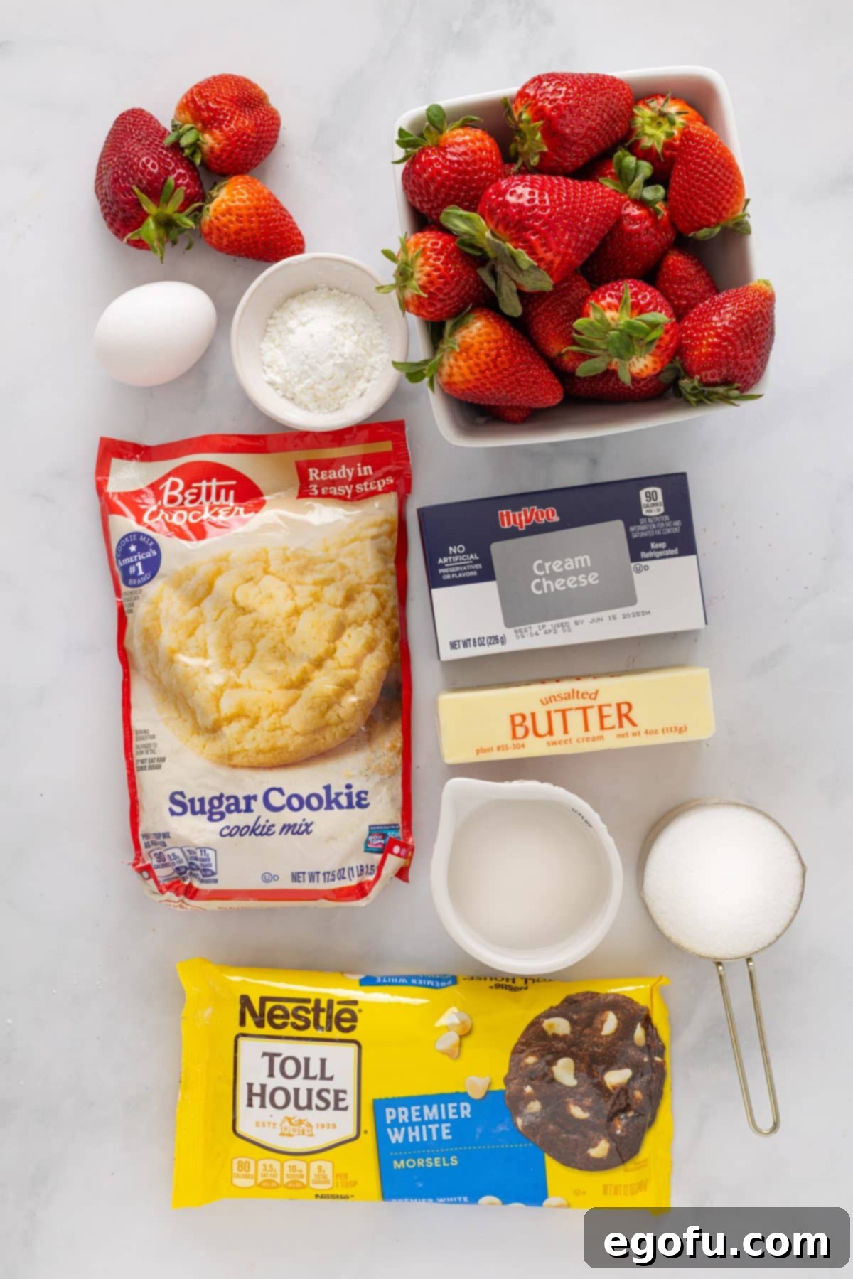 A flat lay image displaying all the ingredients needed: fresh strawberries, eggs, sugar, butter, cream cheese, sugar cookie mix, white chocolate chips, water, and cornstarch, ready for preparation.