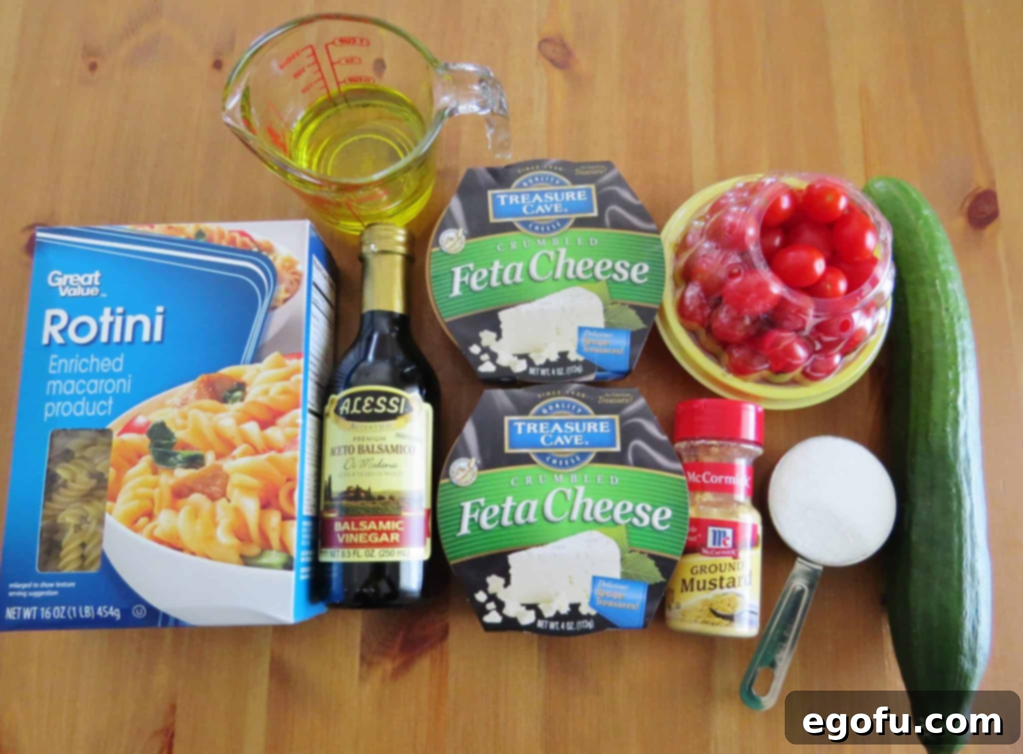 Close-up of fresh ingredients for pasta salad: rotini pasta, crumbled Feta cheese, sliced cucumber, halved cherry tomatoes, diced red onion, and small bowls with sugar, balsamic vinegar, and dry mustard.