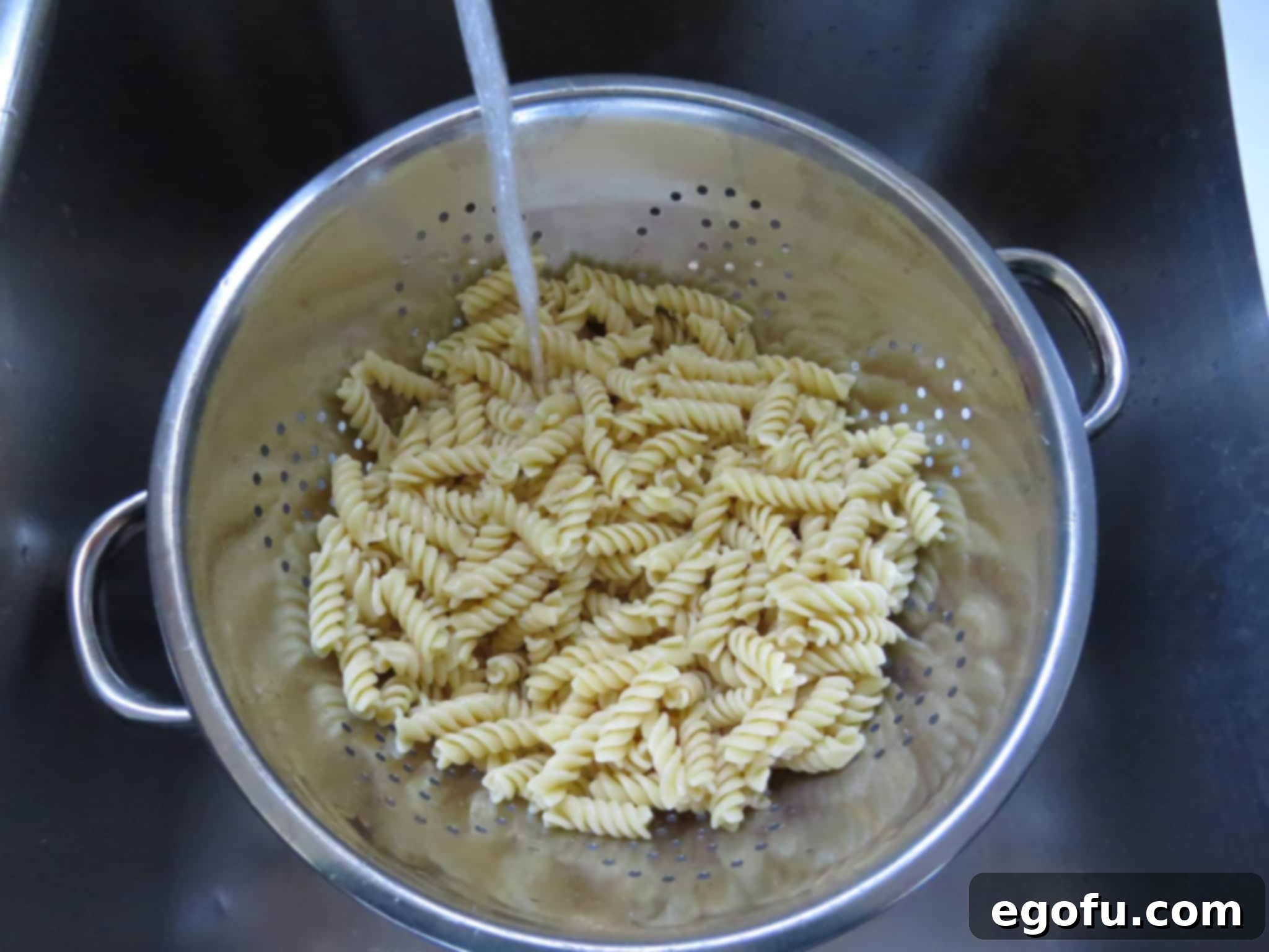 Close-up of cooked rotini pasta being rinsed under cold water in a colander, ready for pasta salad preparation.
