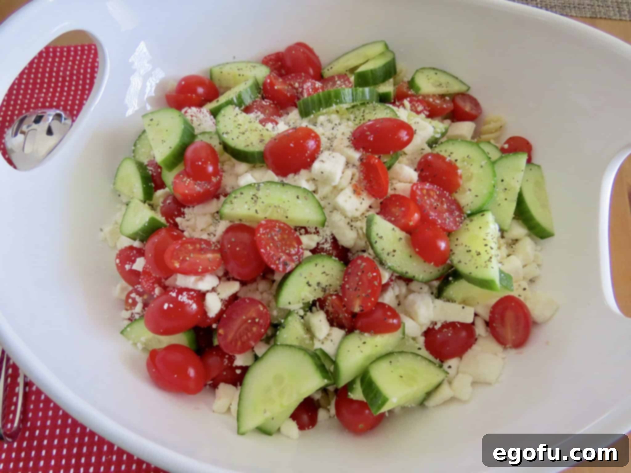 A large white serving bowl filled with a mix of rotini pasta, vibrant cherry tomatoes, crisp cucumbers, and crumbled feta cheese, before dressing.
