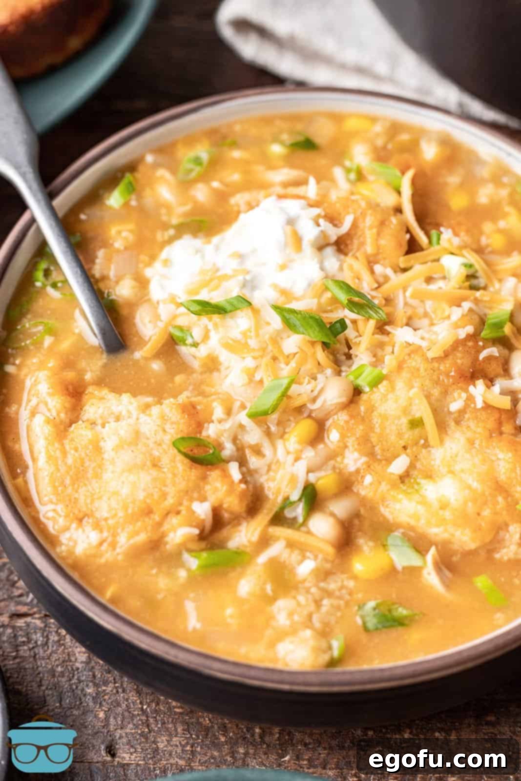 A closeup photo of a bowl of white chicken chili with cornbread dumplings, garnished with a dollop of sour cream and a spoon. 