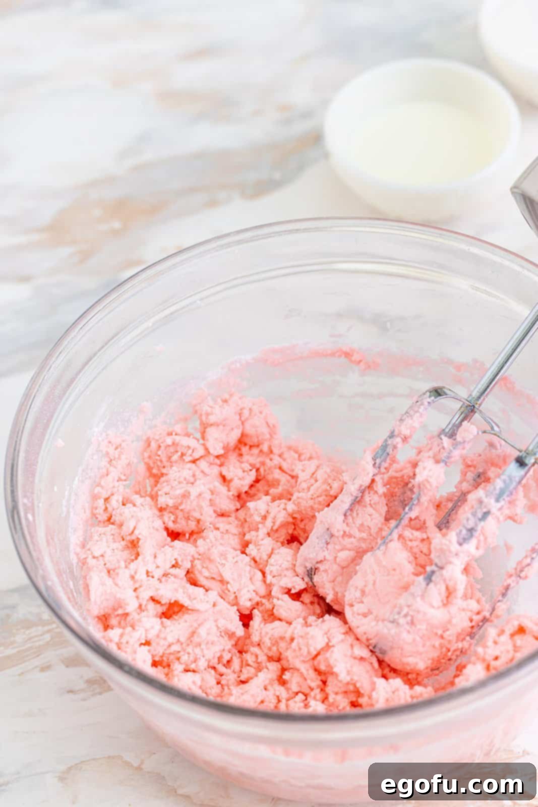 Refreshing Finales 7 Electric hand mixer blending the cream cheese, food coloring, and powdered sugar mixture in a clear bowl, showing the dough starting to come together.