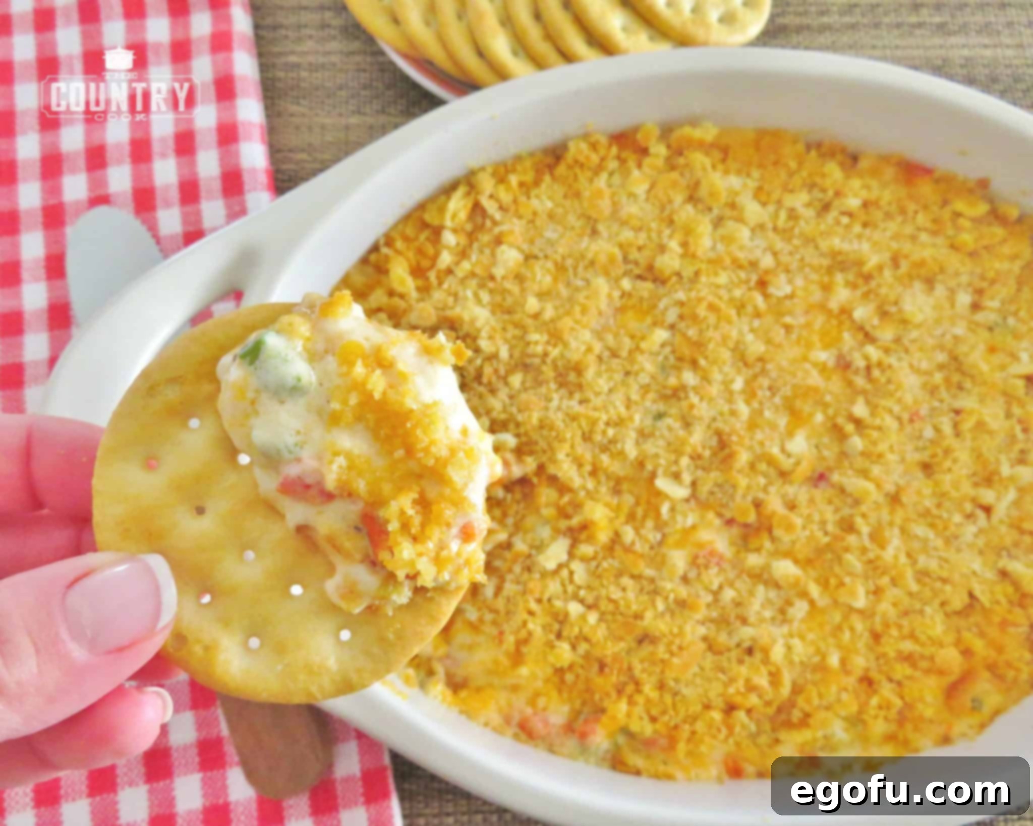 A hand holding a buttery cracker topped with warm pimiento cheese dip, showcasing its creamy texture.