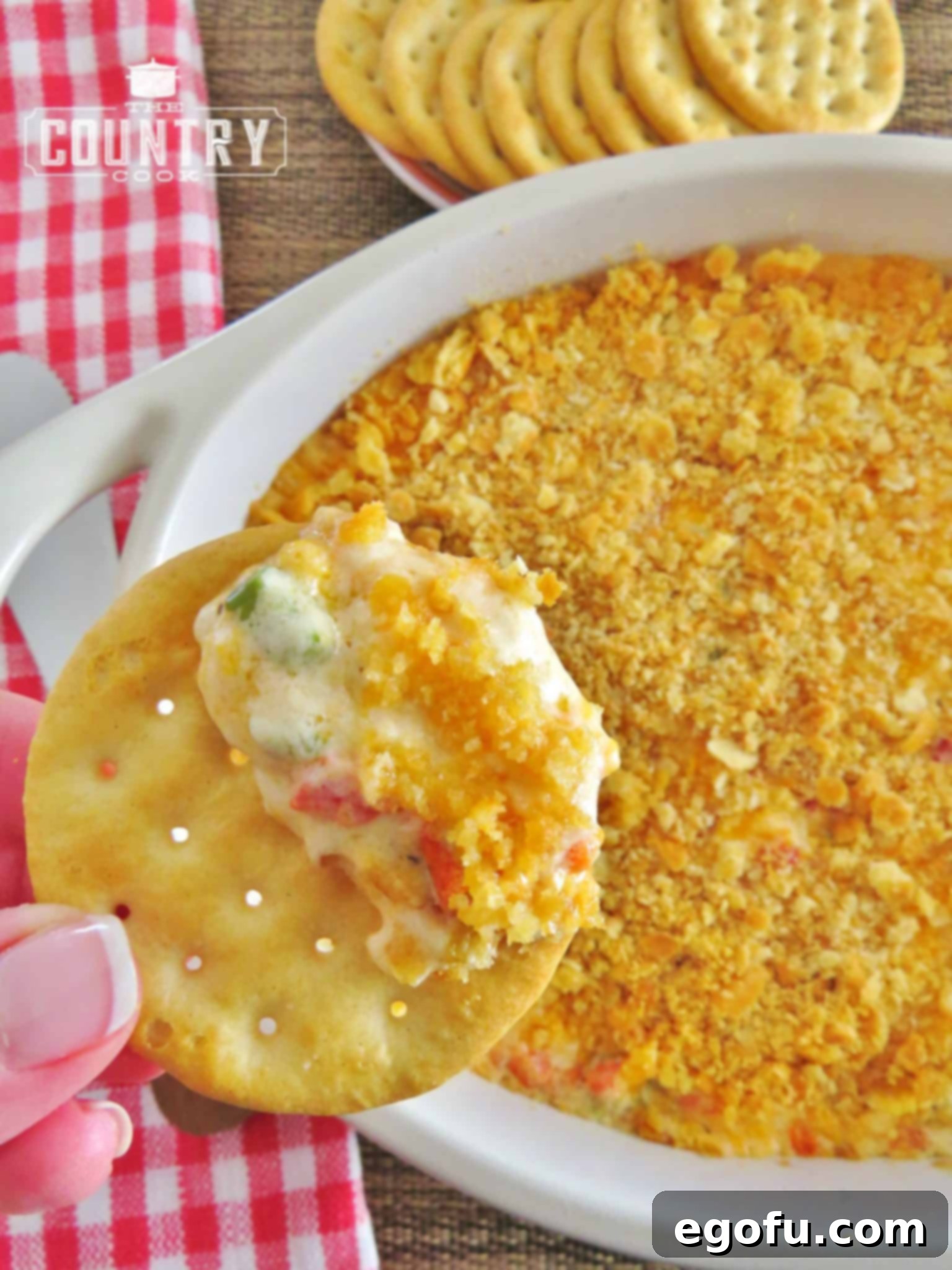 An overhead photo of a hand scooping warm pimiento cheese dip onto a butter cracker, highlighting its tempting presentation.
