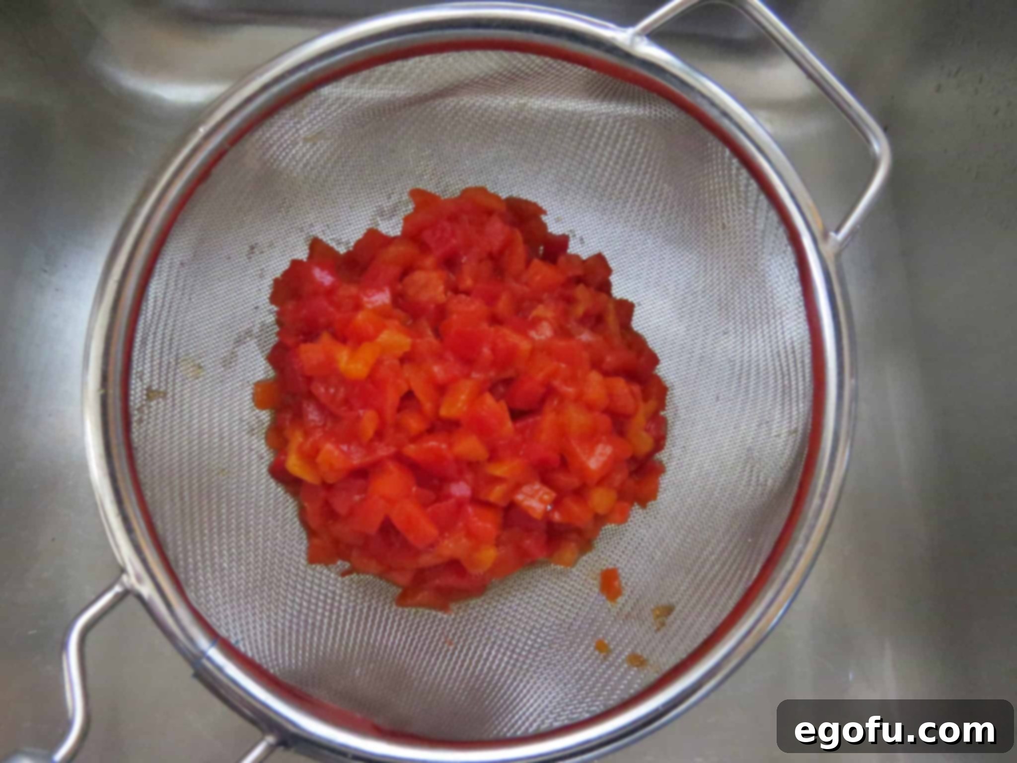 A jar of diced pimientos being thoroughly drained in a metal mesh strainer, emphasizing the importance of removing excess liquid.