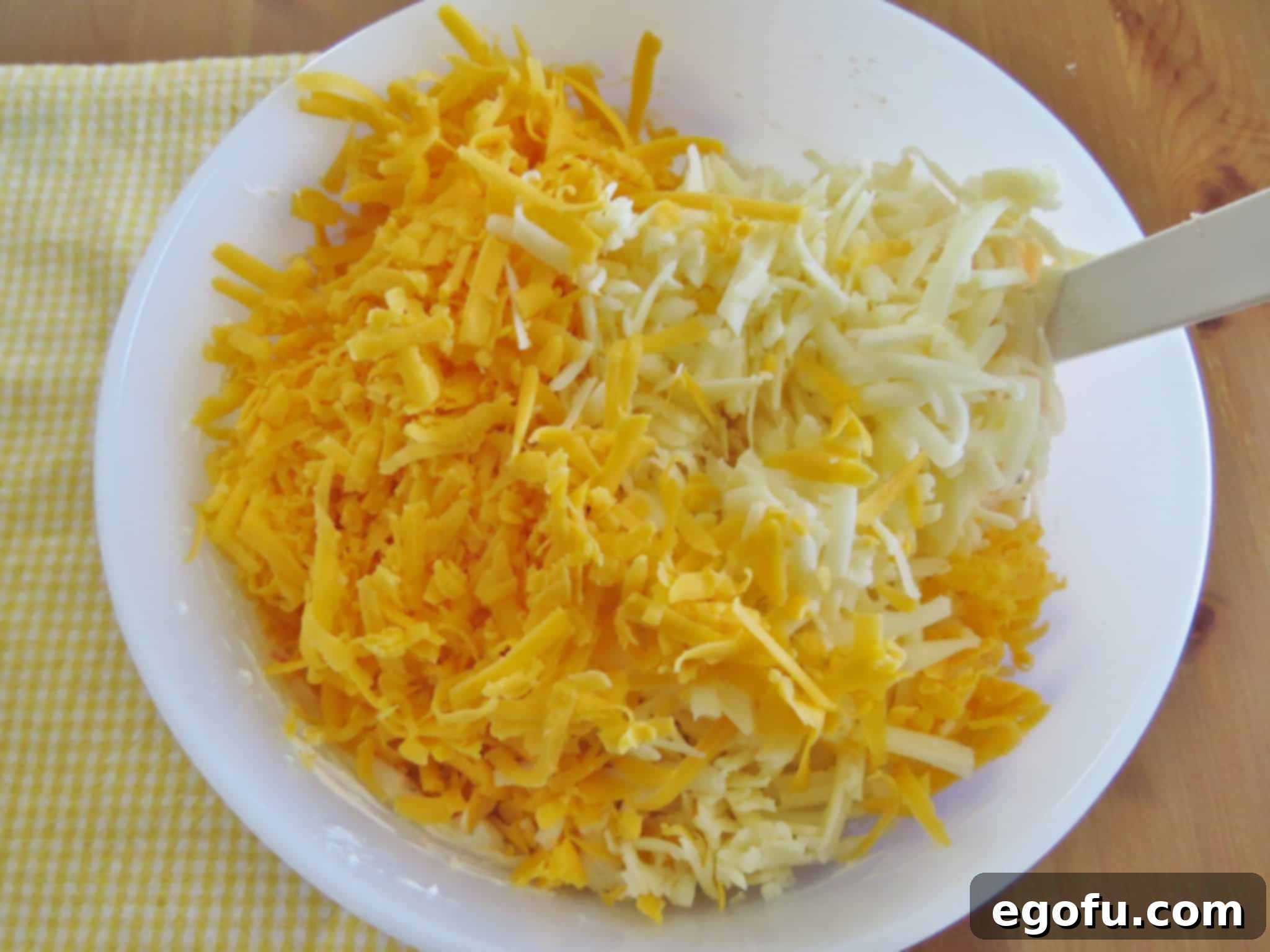 Shredded cheddar cheese being added to the rich, seasoned cream cheese mixture in a large bowl, ready for final mixing.