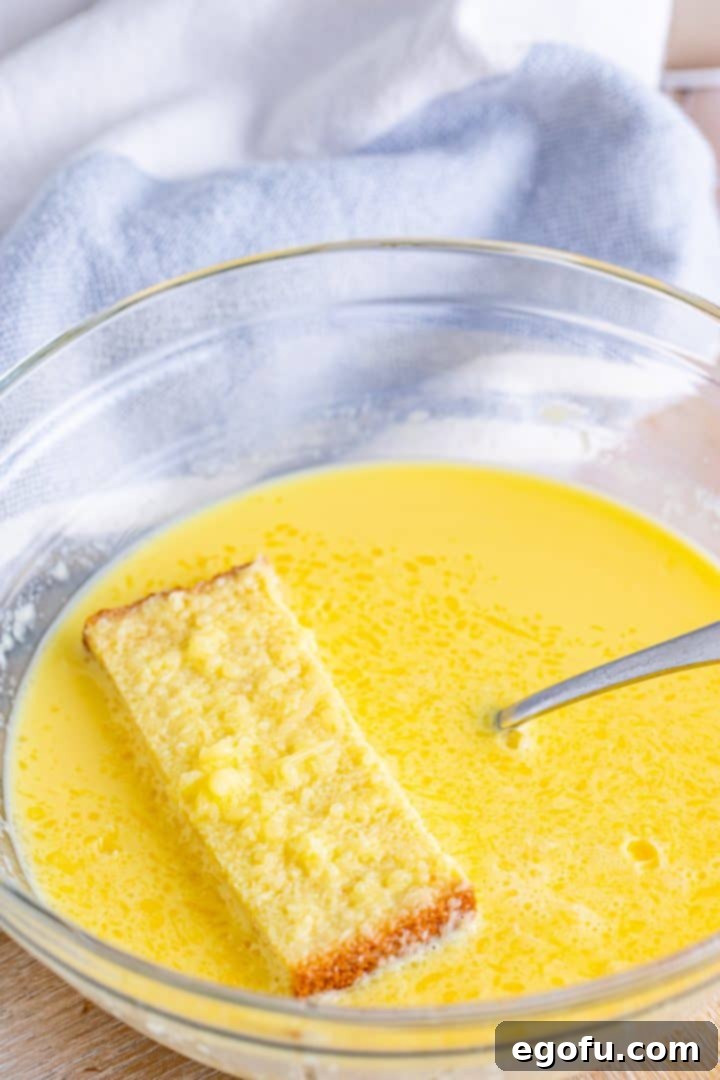 A small, thick slice of white bread is gently dipped into the golden egg mixture in a glass bowl, ensuring an even coating on both sides.