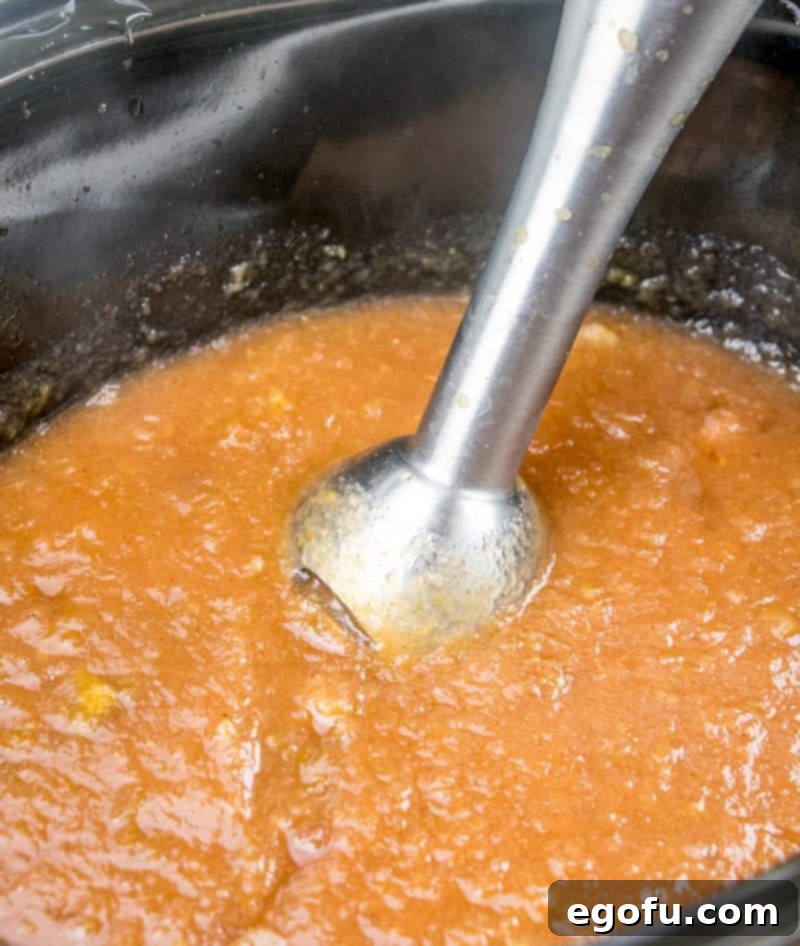 An immersion blender is used to gently chop cooked apples in a crock pot, blending the skins into the sauce.