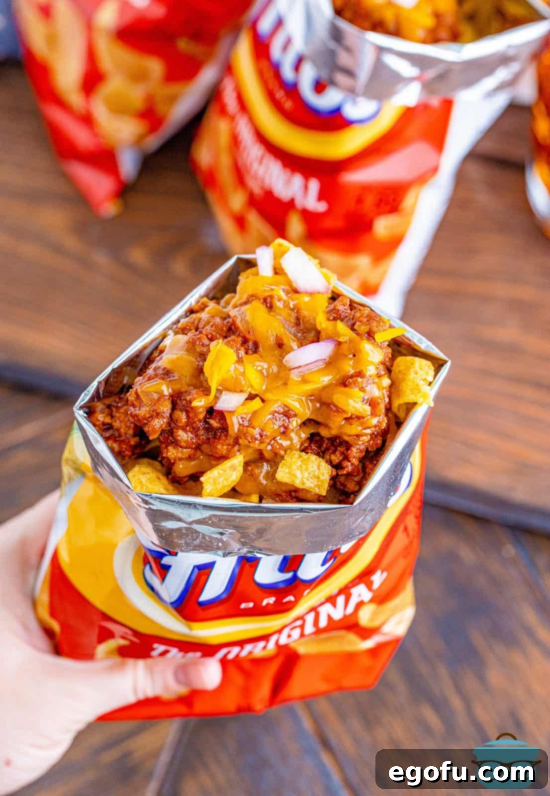 a hand holding a bag of Fritos corn chips that is topped with chili, cheese and diced onions.