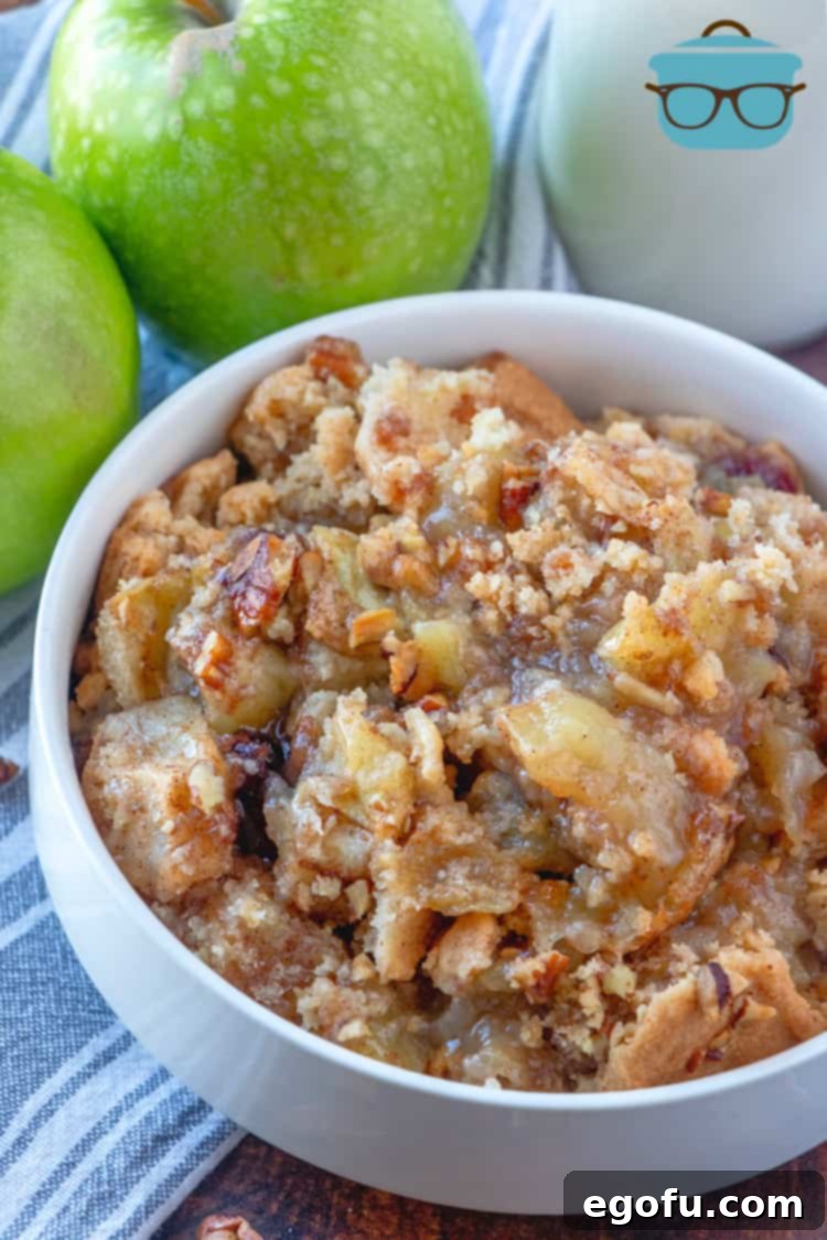 A serving of apple cobbler in a white bowl, with two fresh green apples and a glass of milk in the background, showcasing its rustic charm.