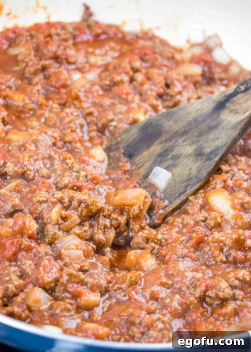 Crushed tomatoes and tomato sauce being stirred into browned ground beef and onions in a large skillet with a wooden spatula.