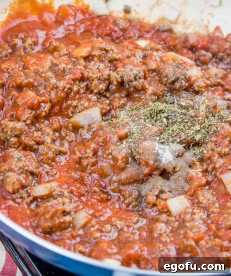 Italian seasoning being sprinkled into the simmering mixture of ground beef, onion, garlic, tomato sauce, and crushed tomatoes in a pot.