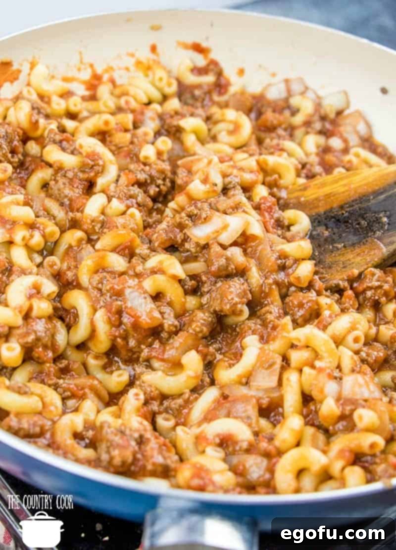 Cooked elbow macaroni noodles being added to the rich tomato sauce with ground beef, diced onion, garlic, and Italian seasoning in a large skillet, ready to be stirred.