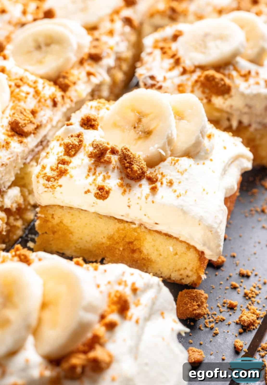 New Orleans Bananas Foster Poke Cake 2 Slices of Bananas Foster Poke Cake on a grey surface, showcasing the layers of cake, caramel, bananas, whipped cream, and Biscoff crumbs.
