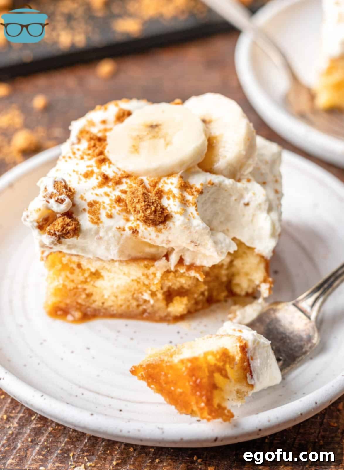 New Orleans Bananas Foster Poke Cake 11 A perfectly sliced piece of Bananas Foster Poke Cake on a small white plate, with a fork holding a bite-sized piece of cake, inviting indulgence.