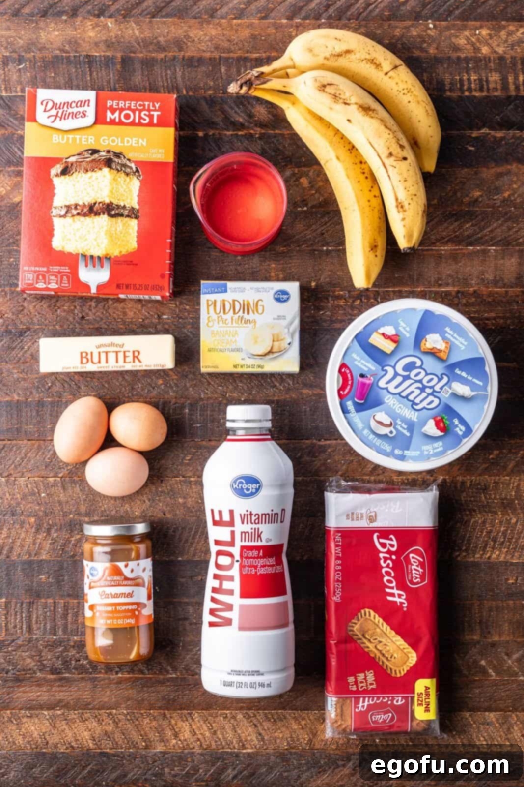 New Orleans Bananas Foster Poke Cake 3 A vibrant display of Bananas Foster Poke Cake ingredients including butter golden cake mix, ripe bananas, instant banana pudding mix, eggs, butter, milk, Cool Whip, caramel sauce, and Biscoff cookies.