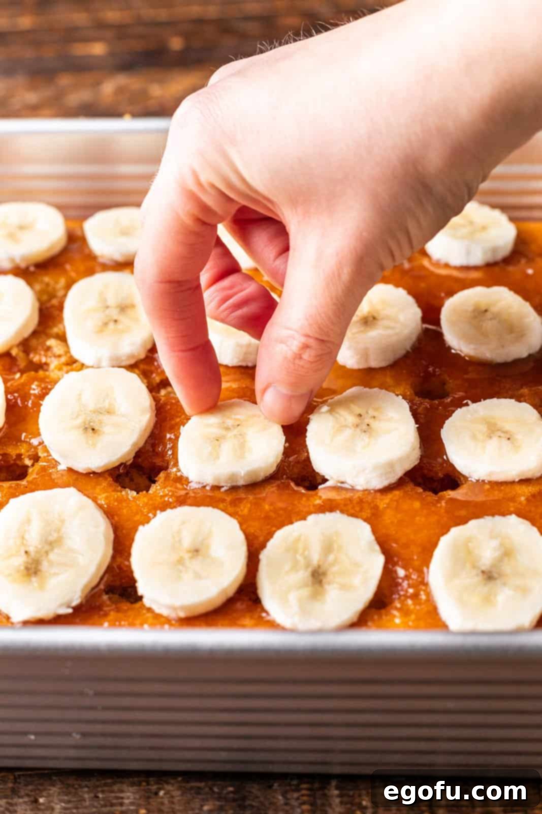 New Orleans Bananas Foster Poke Cake 6 A hand meticulously placing sliced ripe bananas evenly over the caramel-coated cake, creating the next delicious layer.
