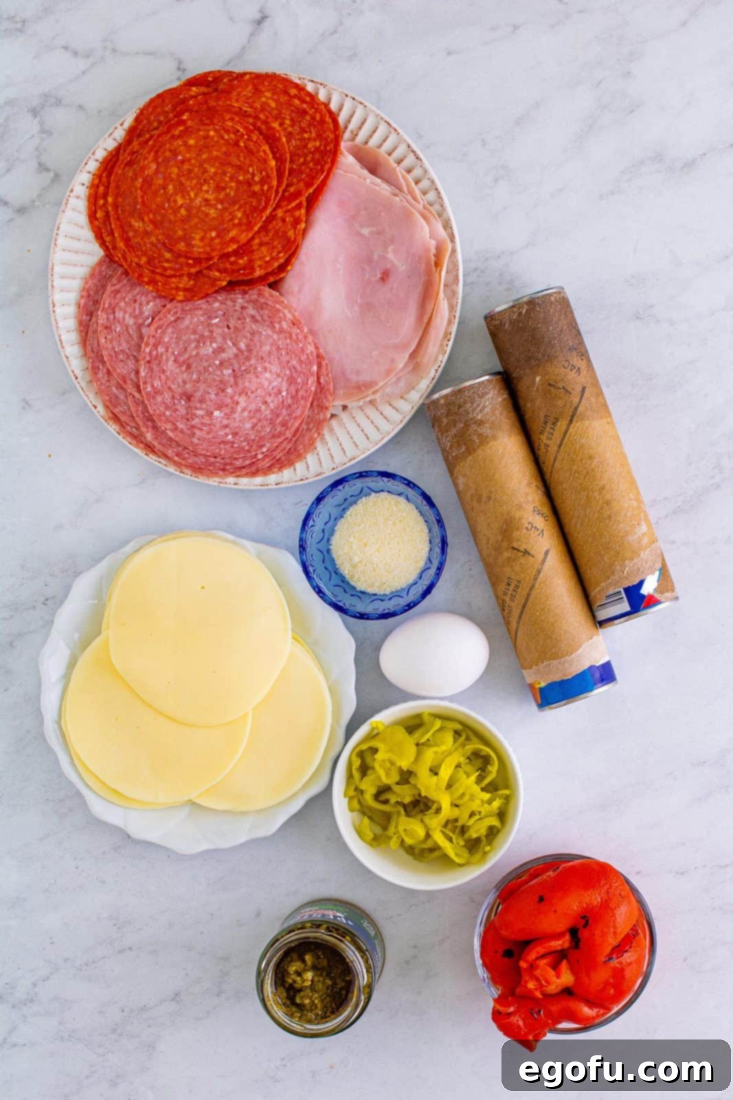 All the essential ingredients laid out: crescent roll dough sheets, provolone cheese, pepperoni, deli ham, Genoa salami, roasted red bell peppers, pepperoncini, egg, pesto, and Parmesan cheese.