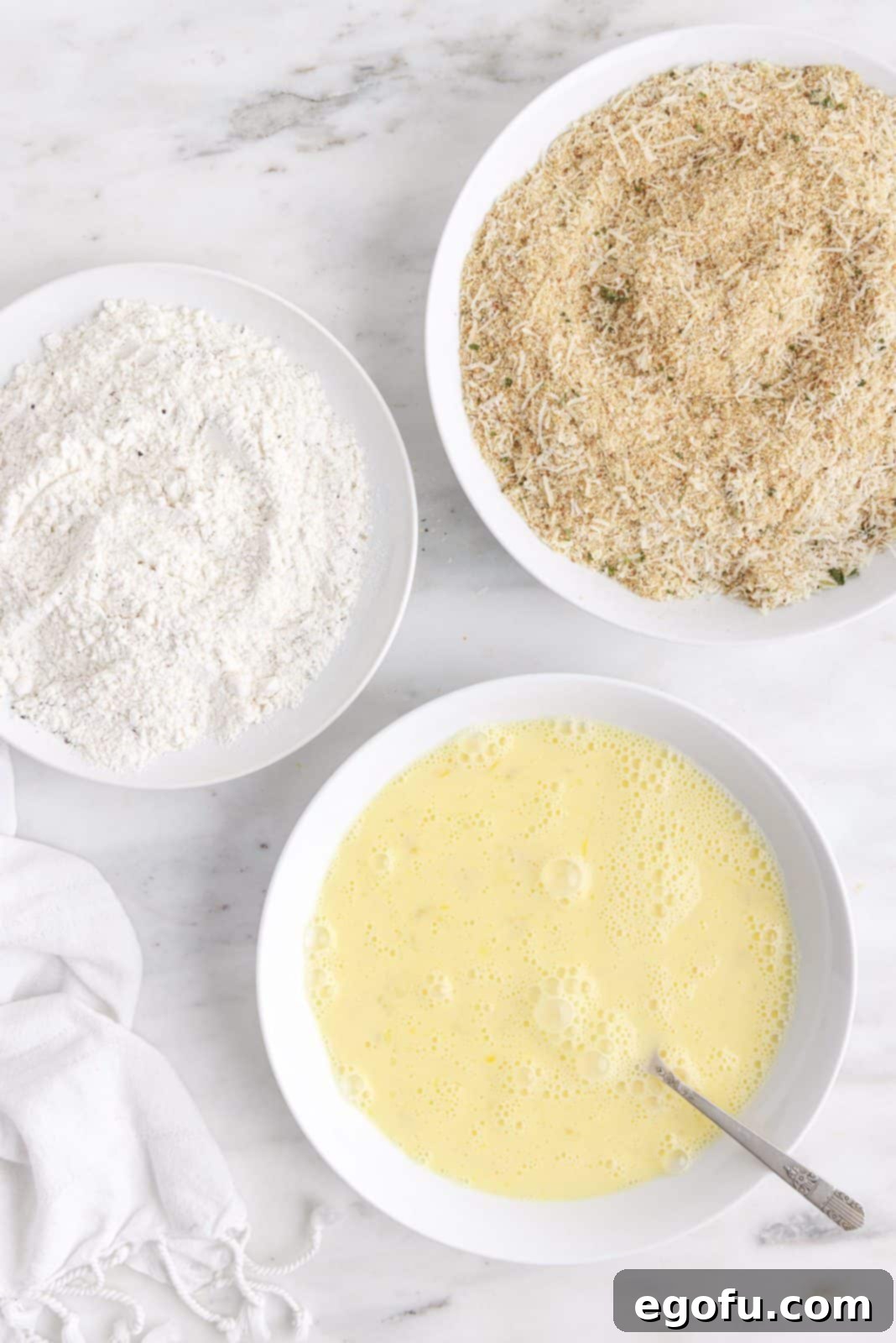 Three shallow dishes set up for dredging: one with flour, salt, and pepper; one with whisked milk and eggs; and one with breadcrumbs and grated cheese, all prepared for coating the lasagna.