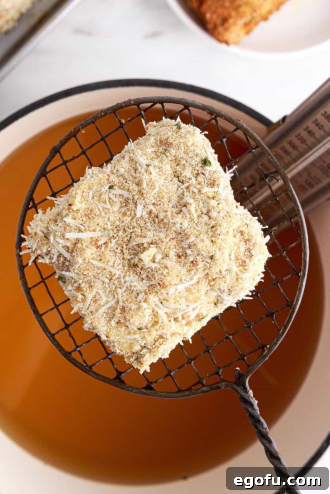 A spider strainer gently lowering a single piece of breaded lasagna into a pot of heated oil, ensuring proper submersion for frying.