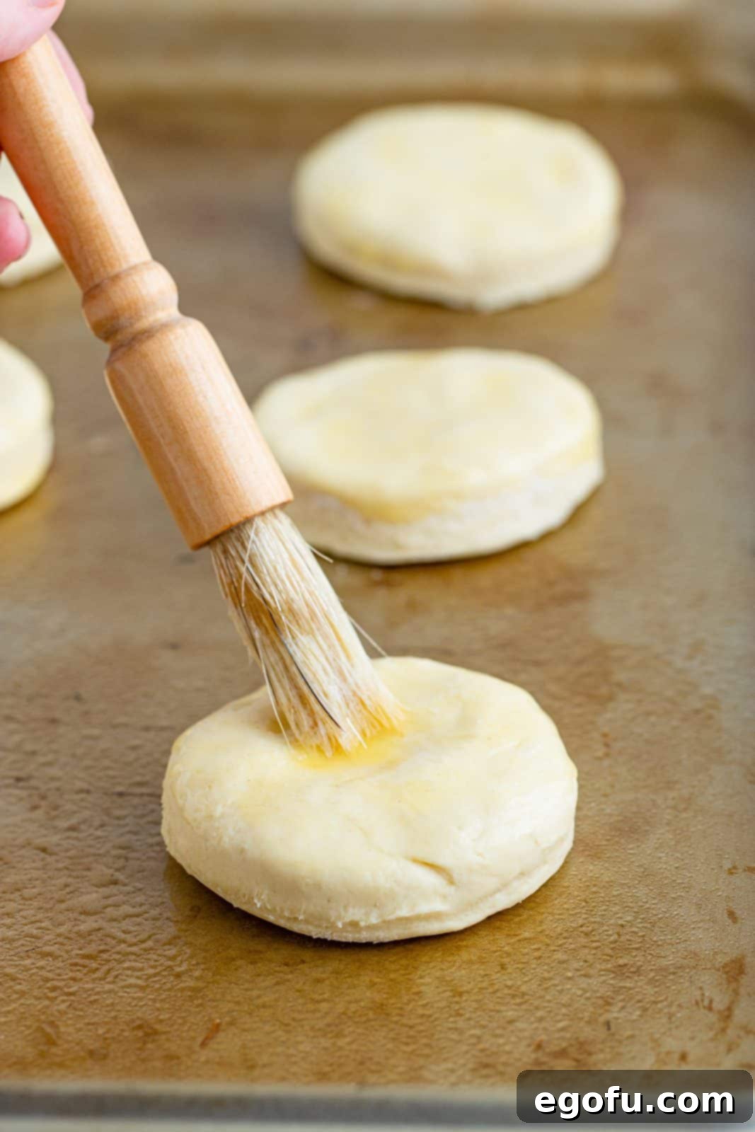 A pastry brush spreading melted butter over the tops of unbaked biscuit dough on a baking sheet.