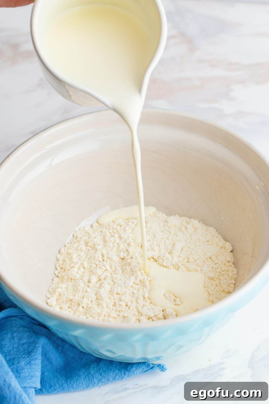 A hand pouring heavy cream from a carton into a white bowl containing self-rising flour.
