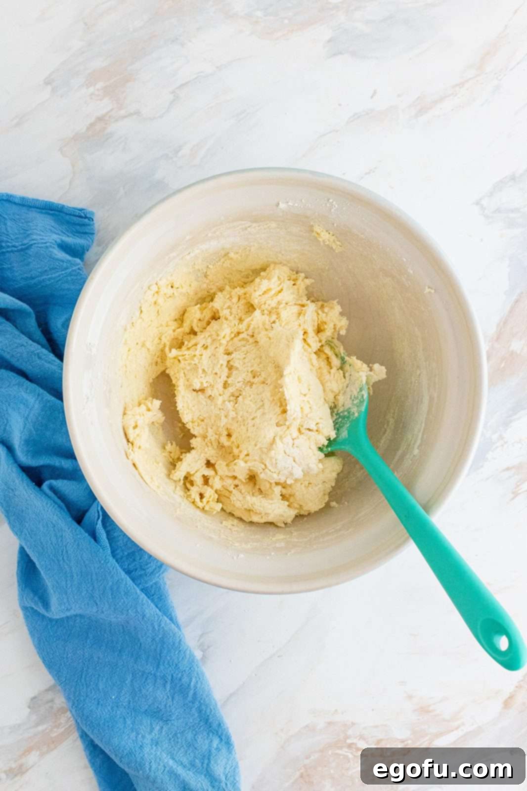 Sticky biscuit dough in a white bowl, being gently mixed with a teal-colored spoonula.