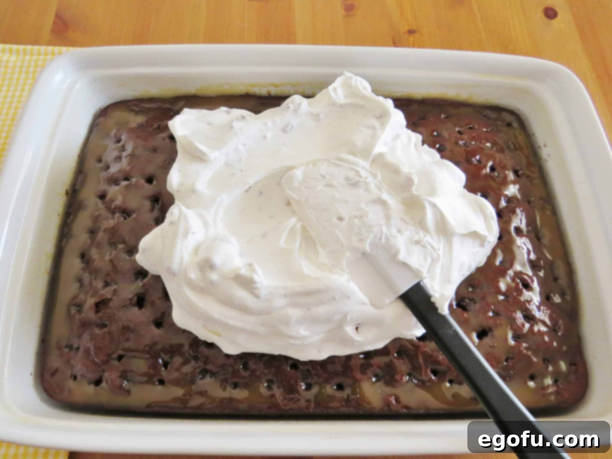 spreading candy bar whipped topping on top of caramel chocolate cake using a white rubber spatula.