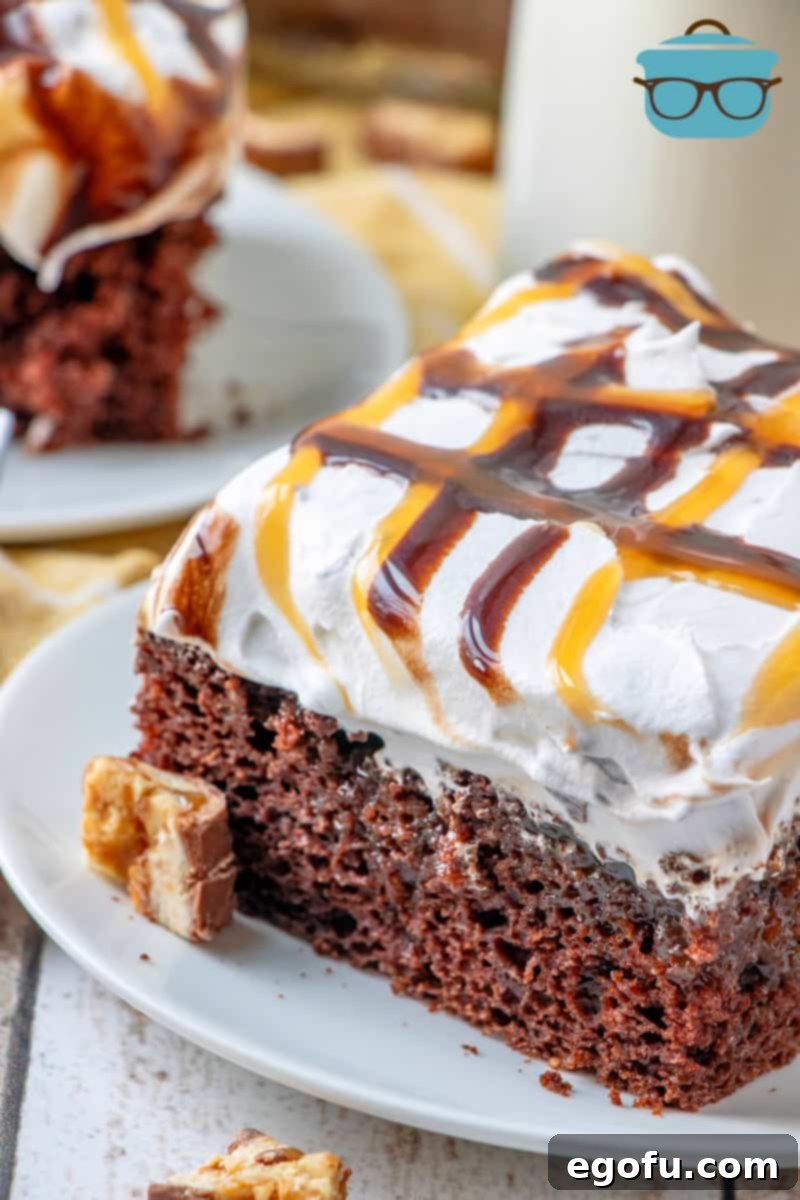slice of Snickers Poke Cake on a small white plate with pieces of chopped Snickers candy bars scattered around.